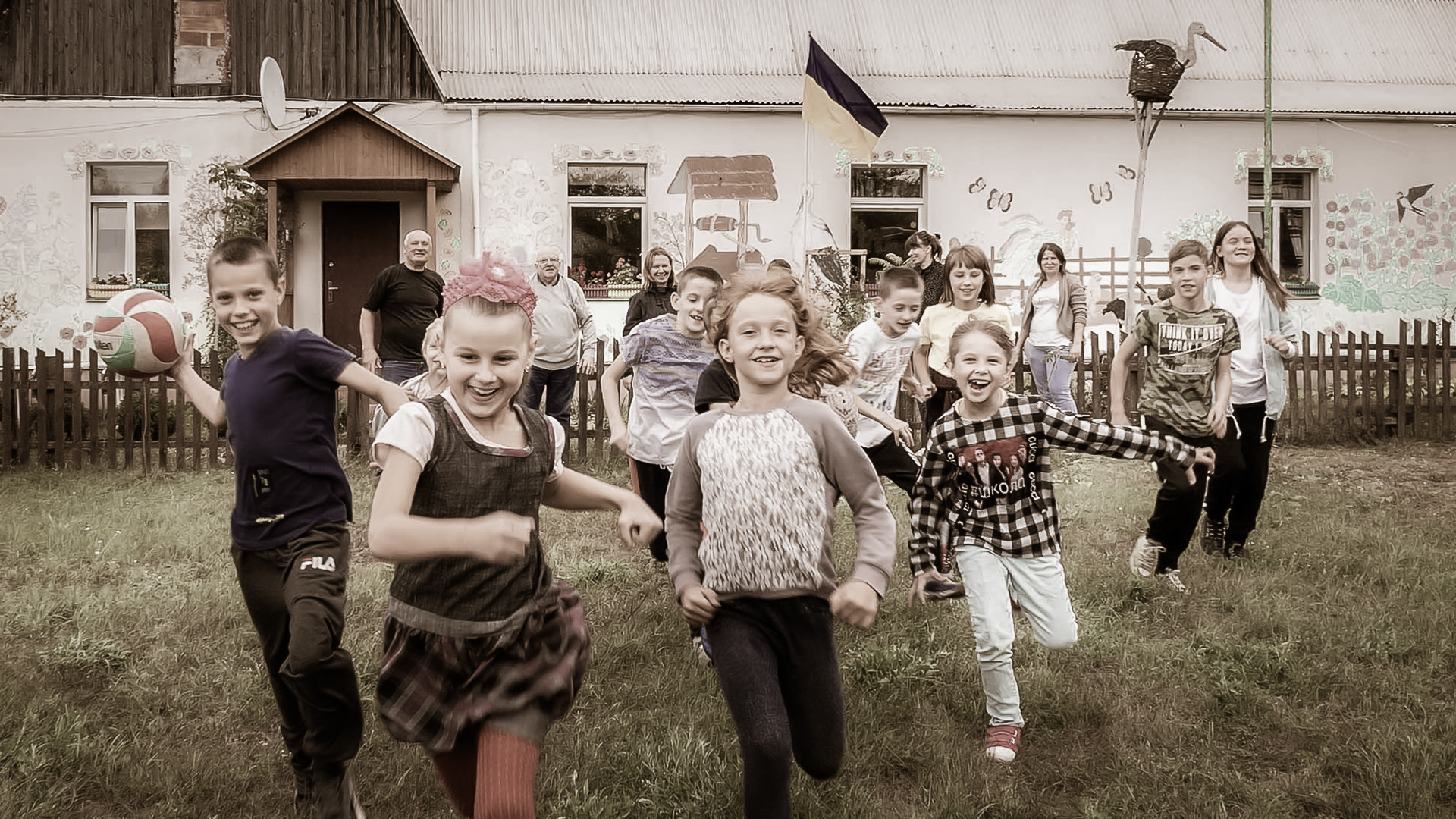 Children run joyfully in the yard of the Frined's house - day center in Ukraine. In the background are smiling adults and the center building. The image conveys hope and togetherness in the midst of war.
